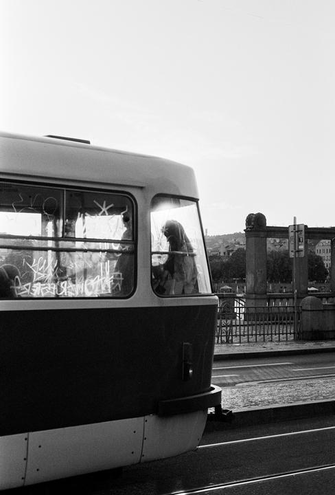 A person is visible through the window of a tram covered in graffiti, with a bridge and buildings in the background.