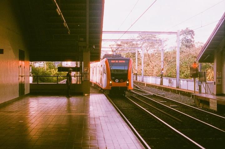 A man stands on a train platform as an orange and white train approaches.
