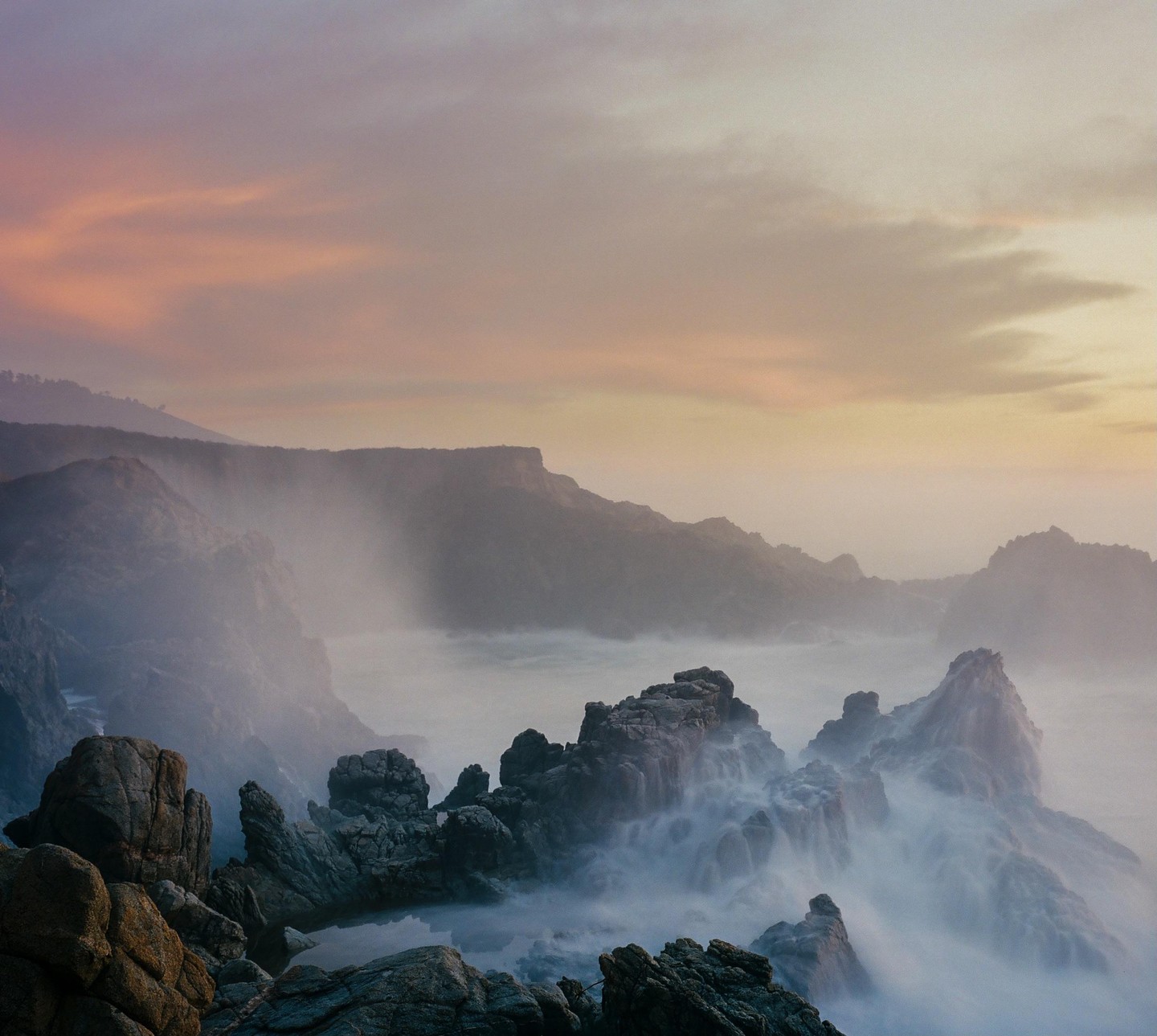 Waves crash against a rocky coastline under a hazy sky at dusk.