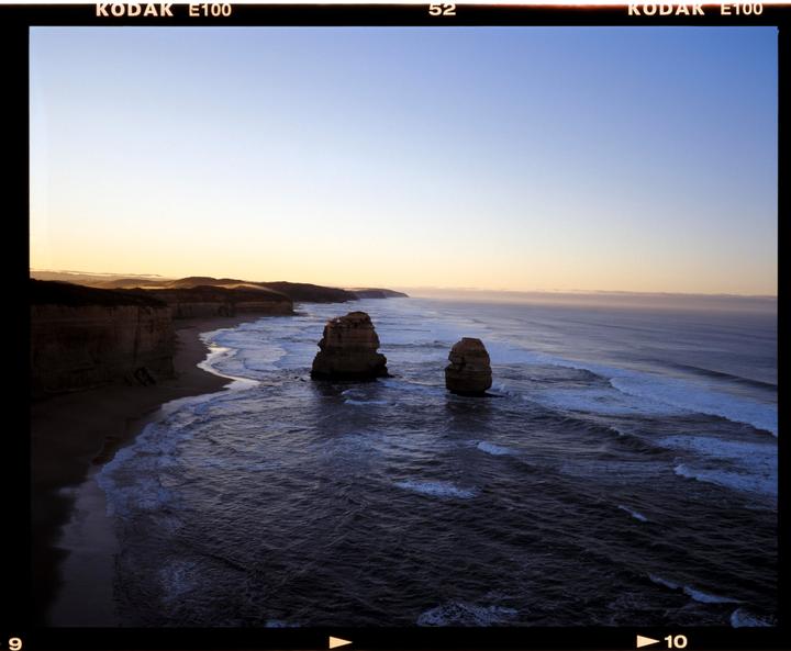 Two large rock formations stand in the ocean near a coastline with cliffs and waves.