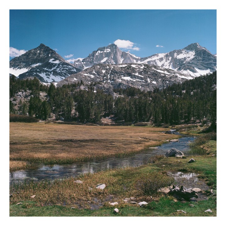 A stream flows through a meadow with snow-capped mountains and a forest in the background.
