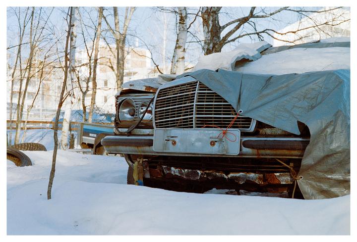A rusty car covered with a tarp is parked in the snow with trees and buildings in the background.