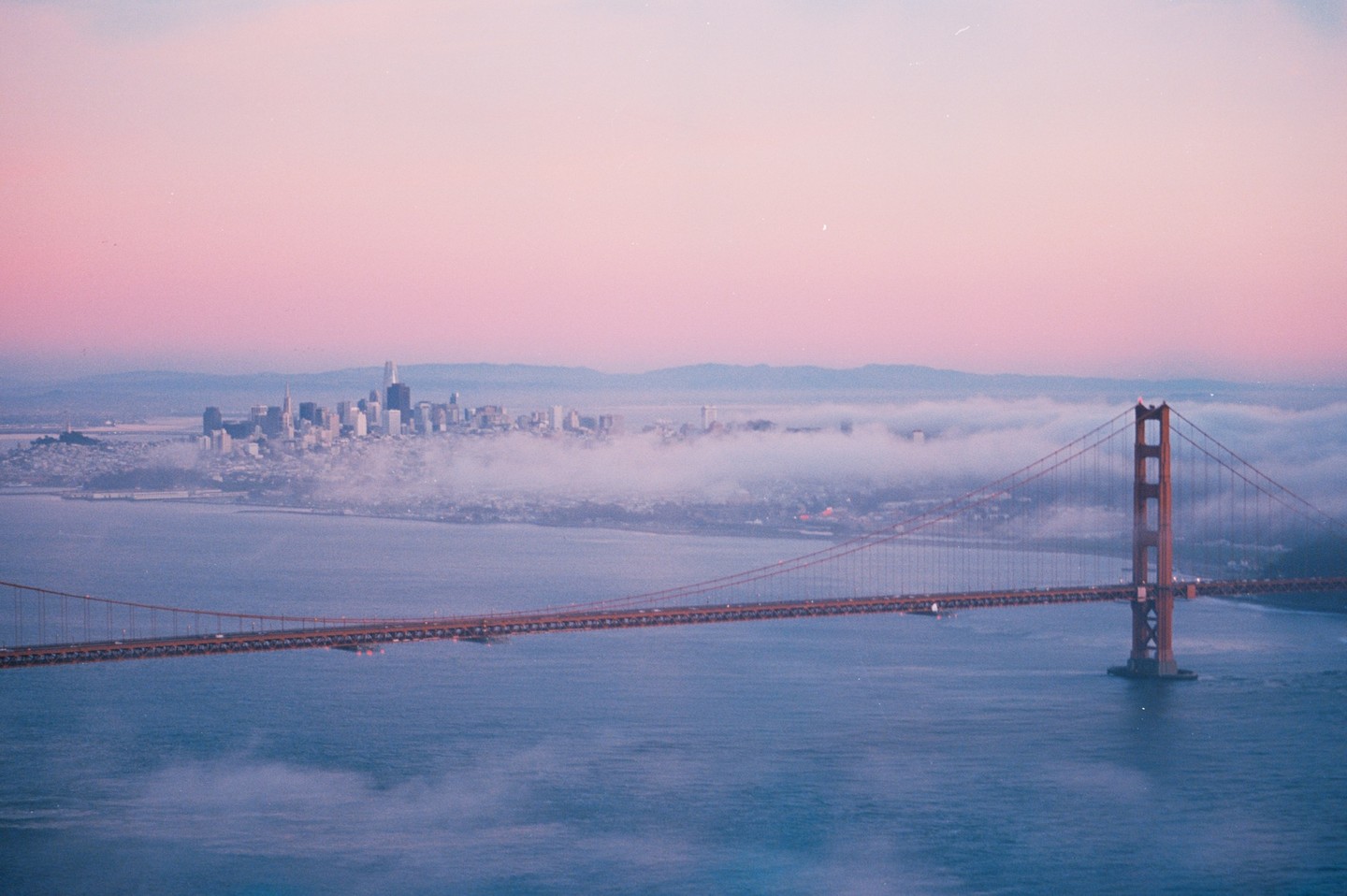 The Golden Gate Bridge spans a body of water with a city skyline in the distance, partially obscured by fog, under a pink and purple sky.