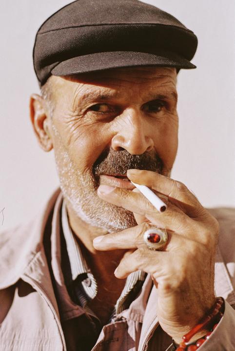 A man with a beard and a cap holds a cigarette to his mouth and wears a ring with a red stone.