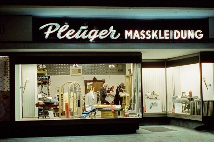 A man reads a newspaper in the window display of a tailor shop at night.