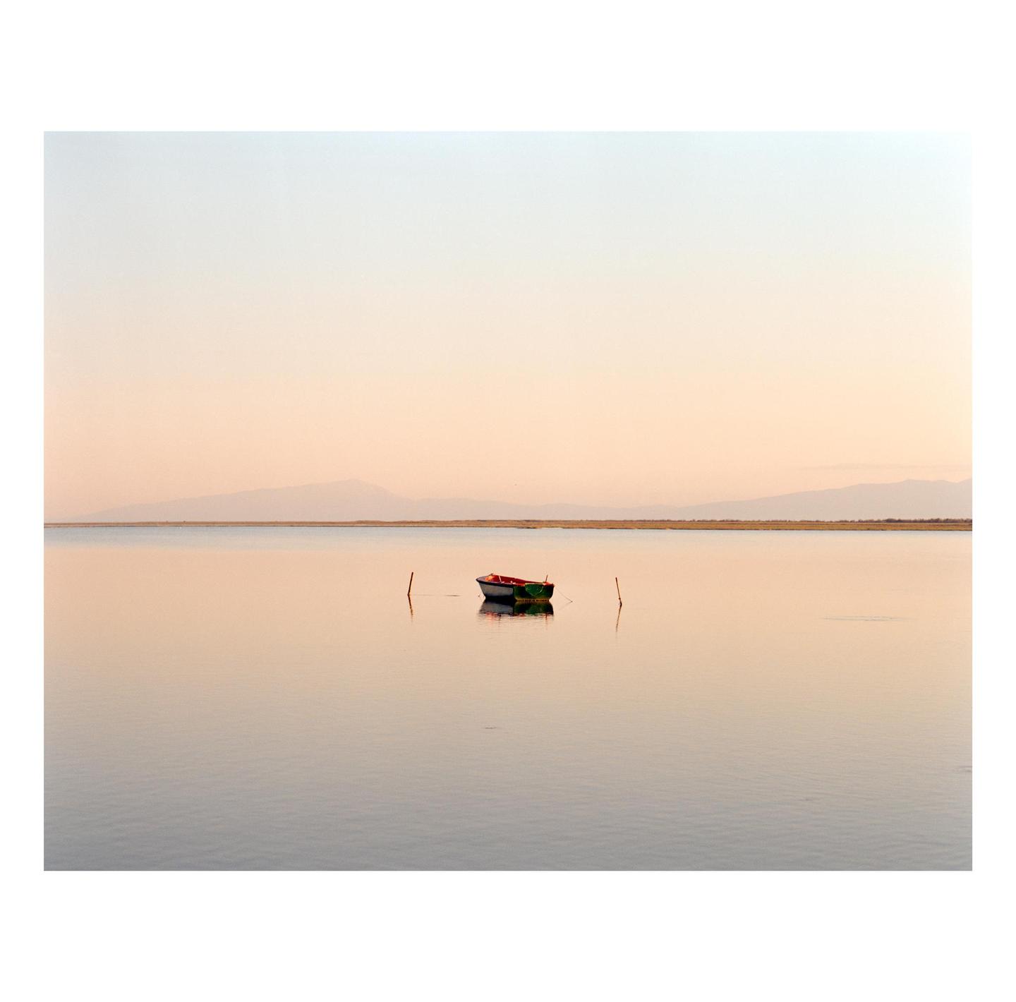 A small boat floats on calm water with mountains in the distance under a pale sky.