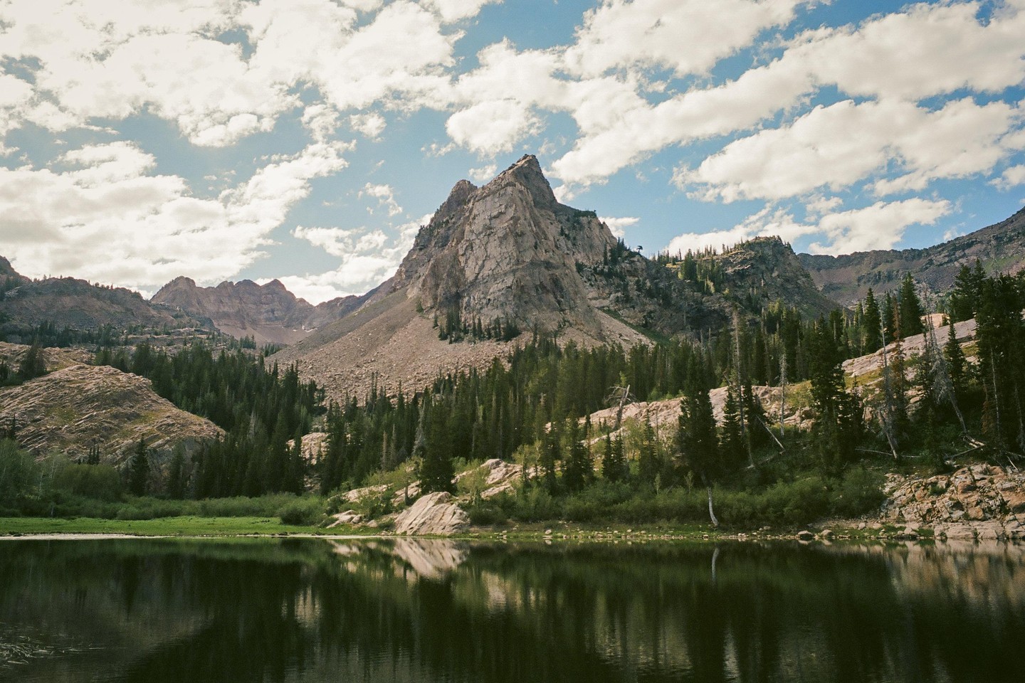 A mountain lake reflects a rocky peak and a forest under a cloudy sky.