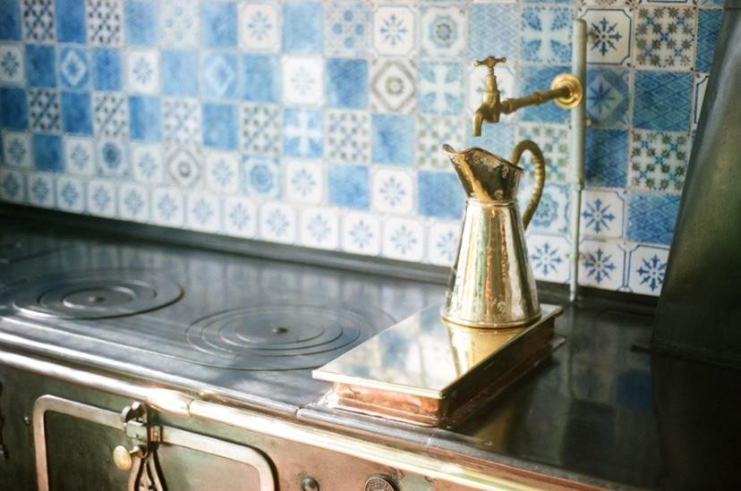 A brass pitcher sits on a stove in front of a tiled wall with a brass faucet.