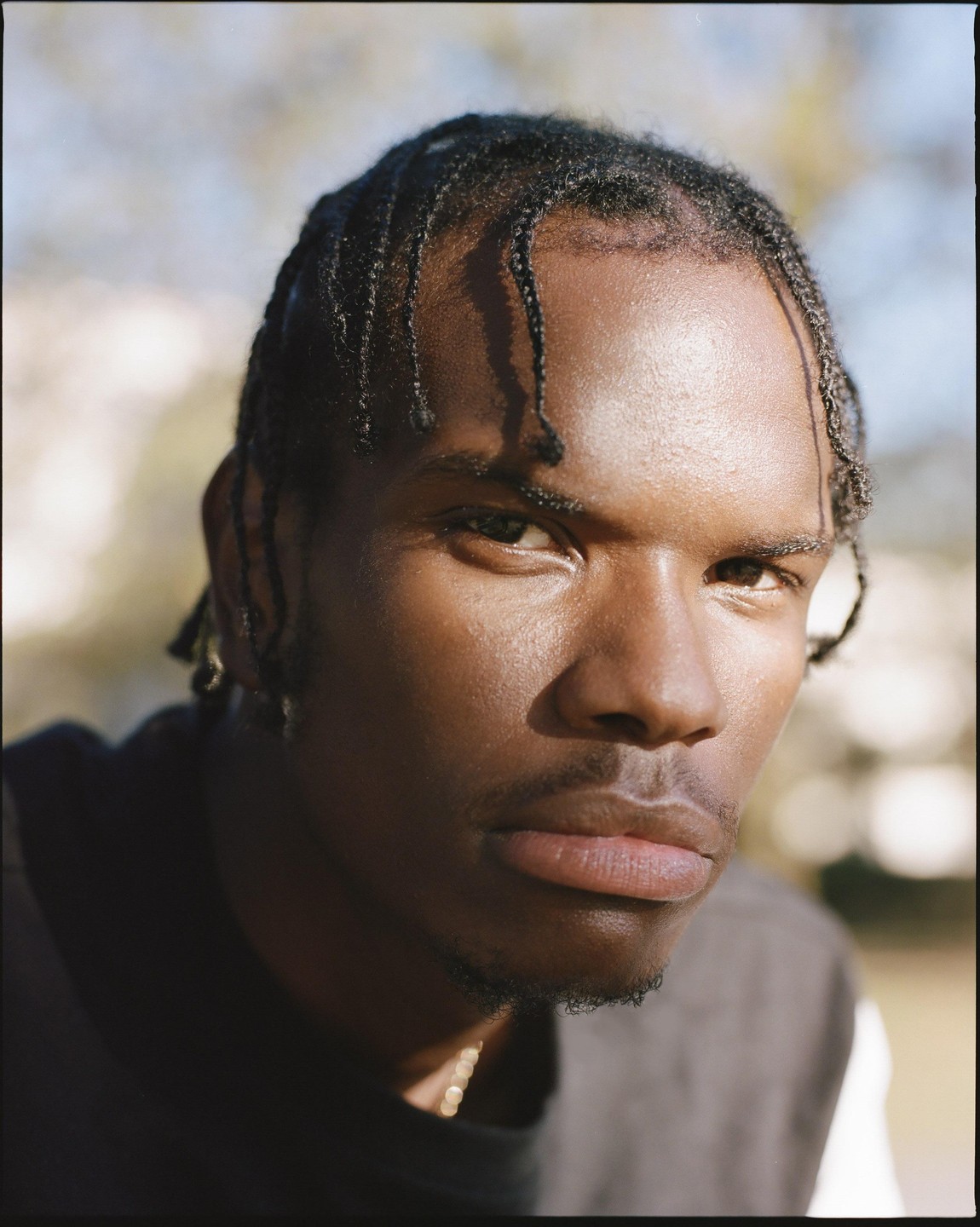 A man with braided hair looks directly at the viewer in a sunlit outdoor portrait.