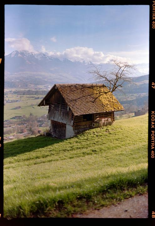 A small wooden cottage with a mossy roof sits on a grassy hill overlooking a valley and mountains.