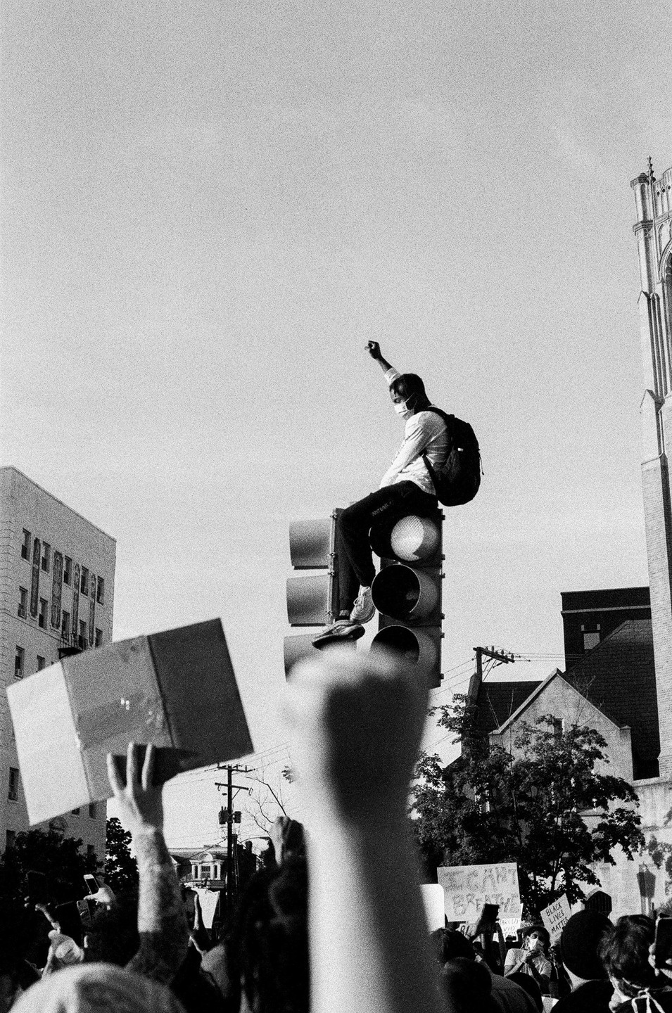 A man wearing a face mask and backpack sits on a traffic light with his fist raised, surrounded by a crowd of people holding signs and raising their fists.