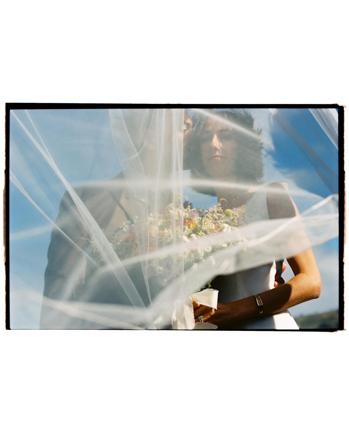 A couple is visible through a wedding veil, with the woman holding a bouquet of flowers.