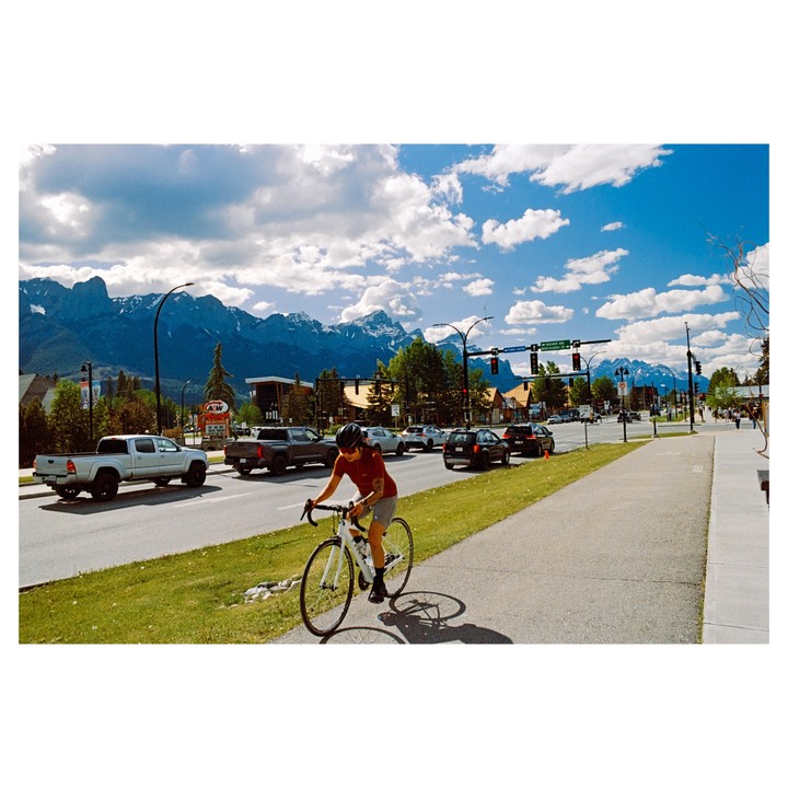 A woman rides a bicycle on a sidewalk next to a street with cars and mountains in the background.