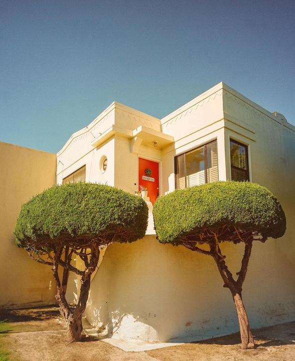 Two manicured trees stand in front of a cream-colored building with a red door.