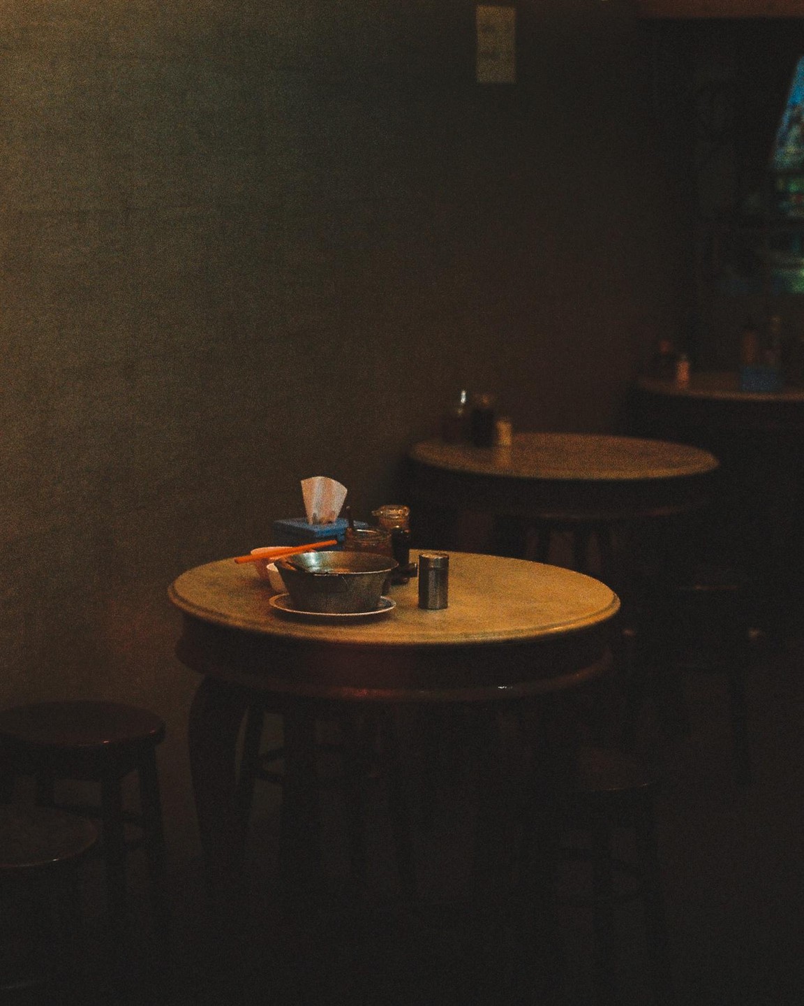 A bowl of food and condiments sit on a round wooden table in a dimly lit restaurant.