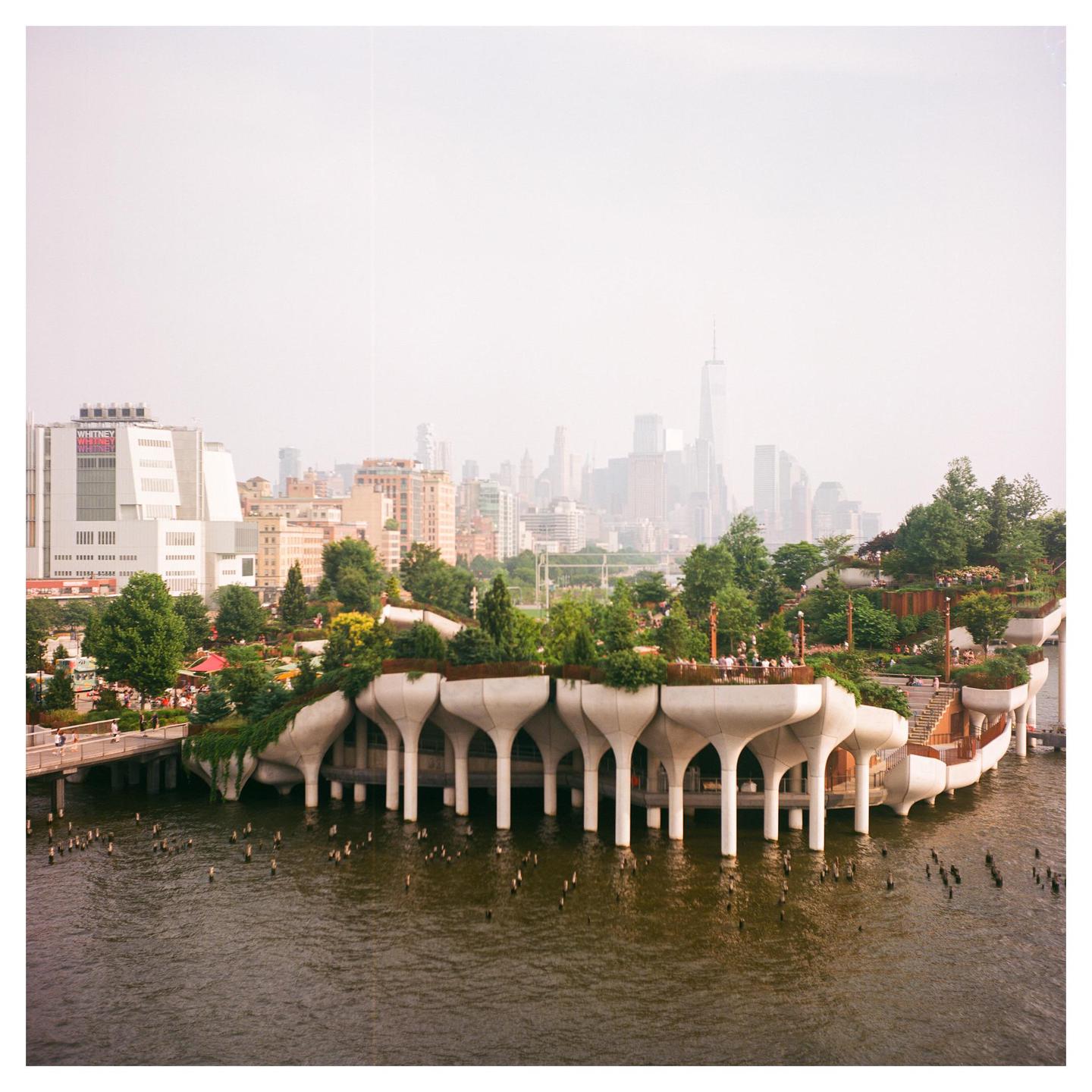 A modern park structure with trees and people on a body of water is visible in the foreground, with a hazy city skyline in the background.