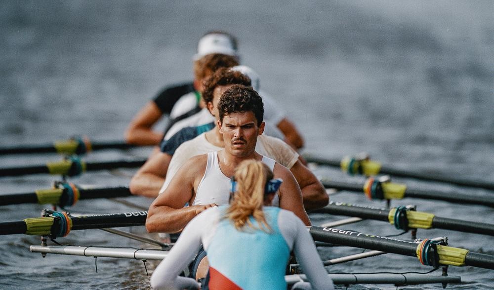 A group of rowers are in a boat on the water, with one man in a white tank top looking forward.