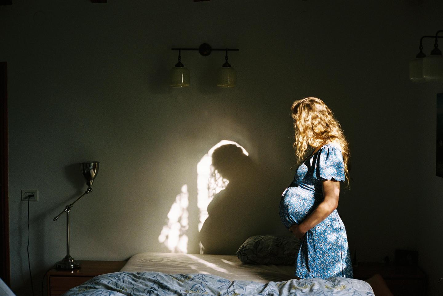 A pregnant woman stands next to a bed, illuminated by a shaft of light that casts her shadow on the wall.