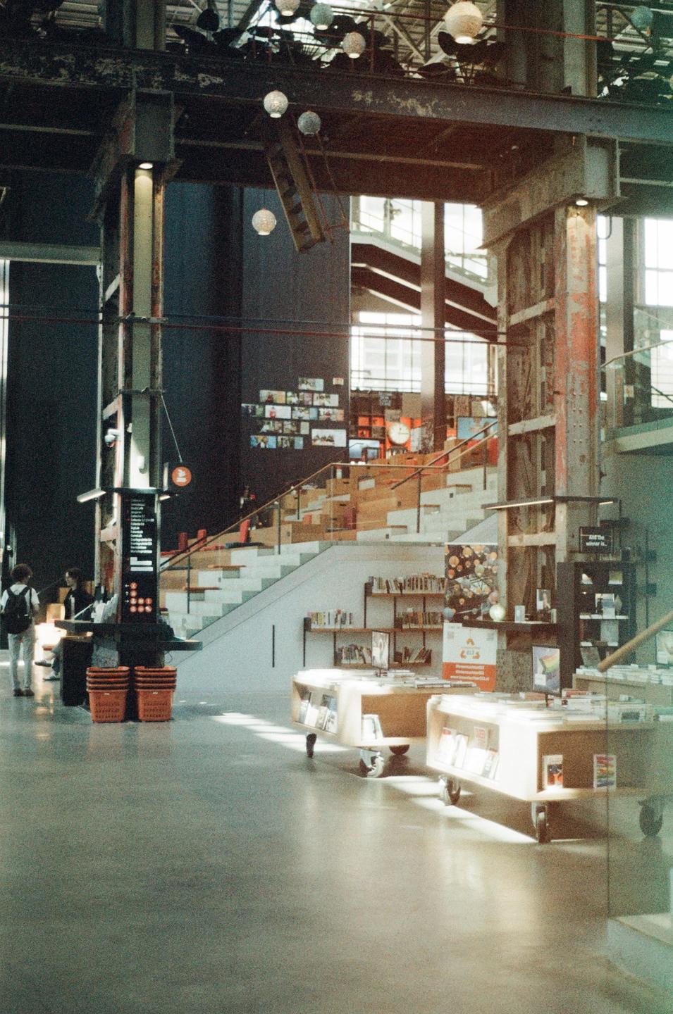 A large, modern library interior features a wide staircase, bookshelves, and people browsing.