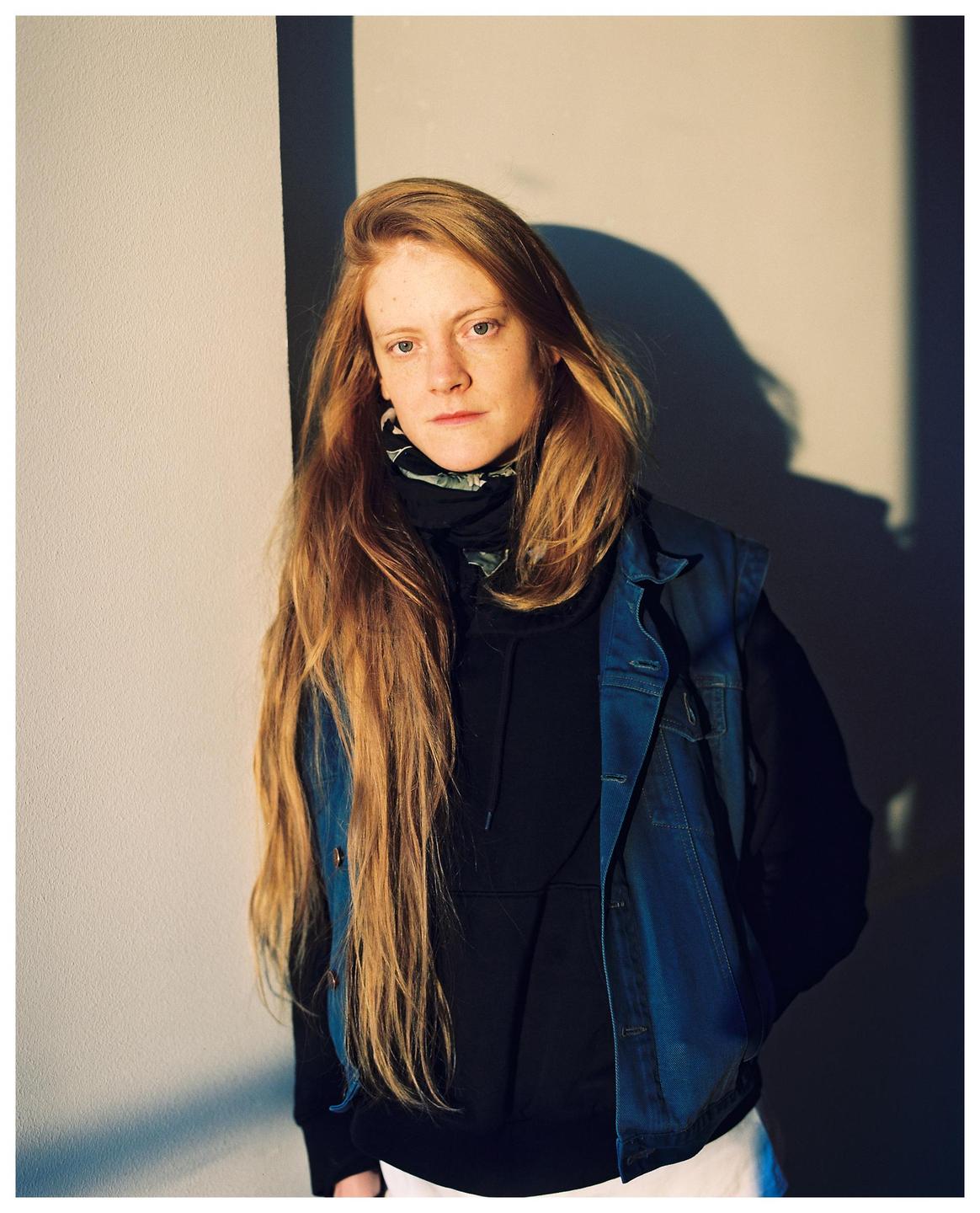A woman with long red hair wears a black hoodie and a denim vest while standing against a wall.
