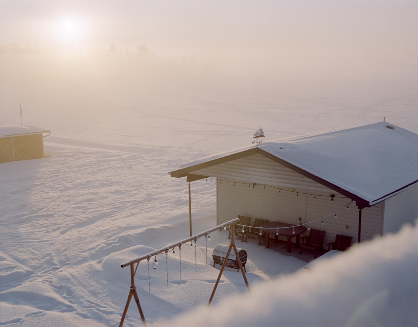 A snow-covered house with a porch and string lights sits in a foggy, winter landscape with another small building in the distance.