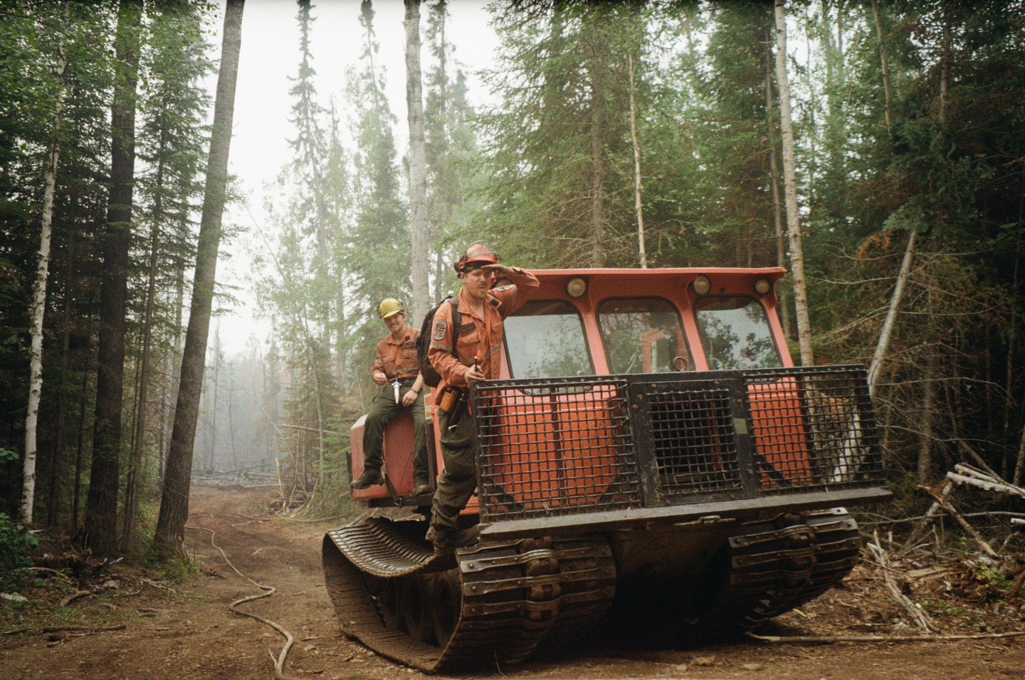 Two men in work uniforms are on a large red tracked vehicle in a forest.