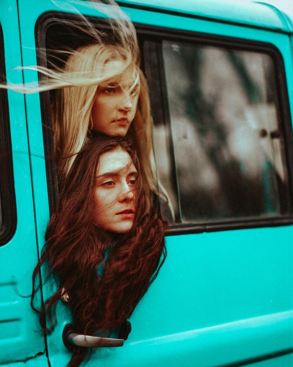 Two women with long hair are looking out of the window of a turquoise vehicle.