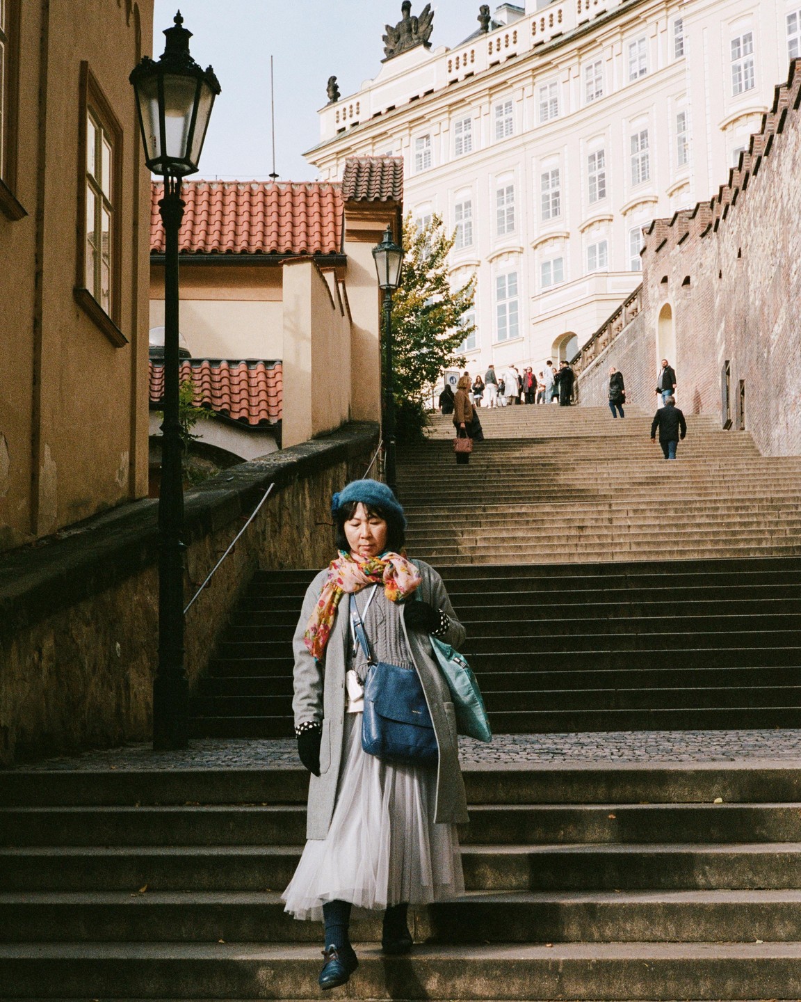 A woman in a blue beret and gray coat walks down a long stone staircase in Prague.