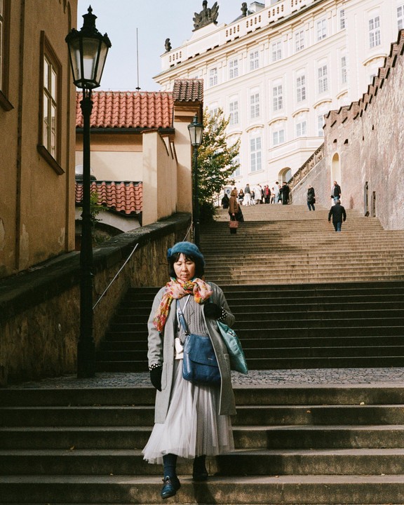 A woman in a blue beret and gray coat walks down a long stone staircase in Prague.
