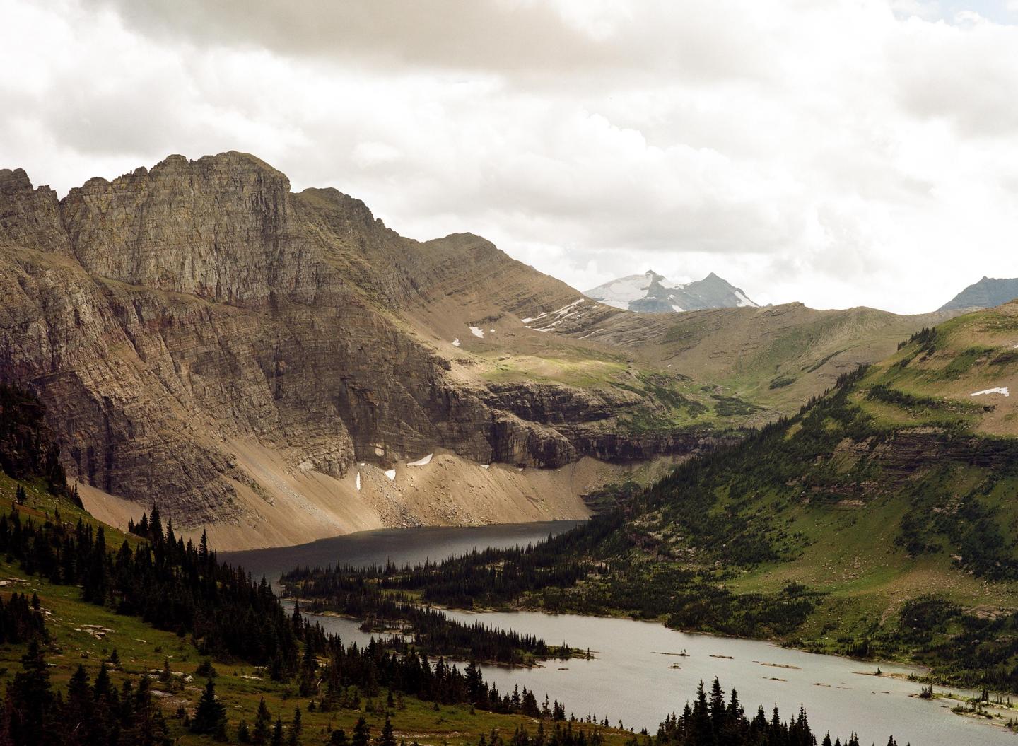 A wide mountain valley with a lake and evergreen trees is visible under a cloudy sky.