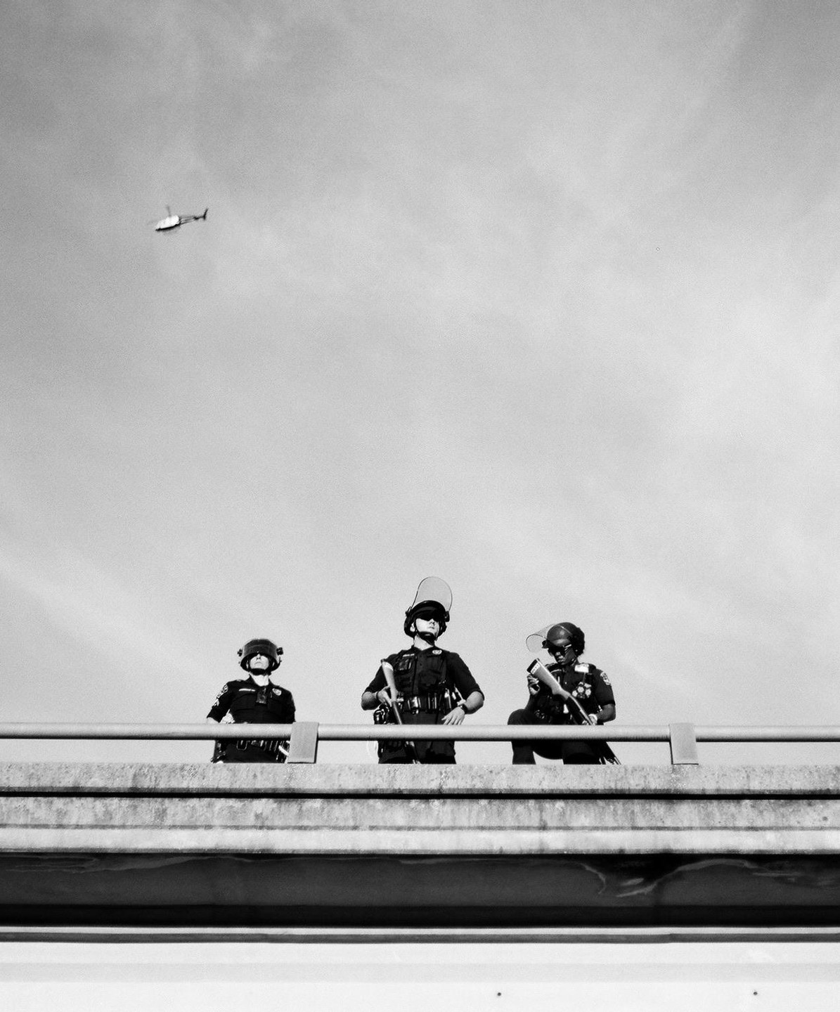 Three police officers in riot gear stand on a bridge with a helicopter flying in the distance.