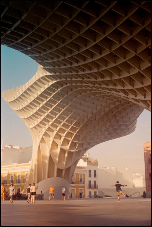 A person balances on one leg in a plaza under a large, modern, gridded structure.