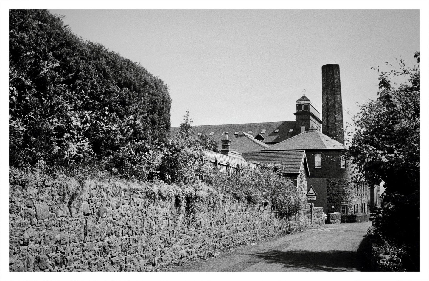 A narrow road runs alongside a stone wall and a tall hedge, leading towards a building with a prominent chimney and a small tower.
