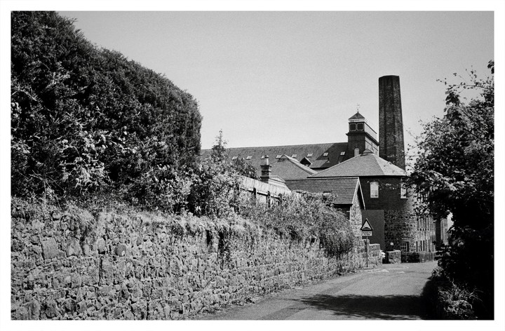 A narrow road runs alongside a stone wall and a tall hedge, leading towards a building with a prominent chimney and a small tower.