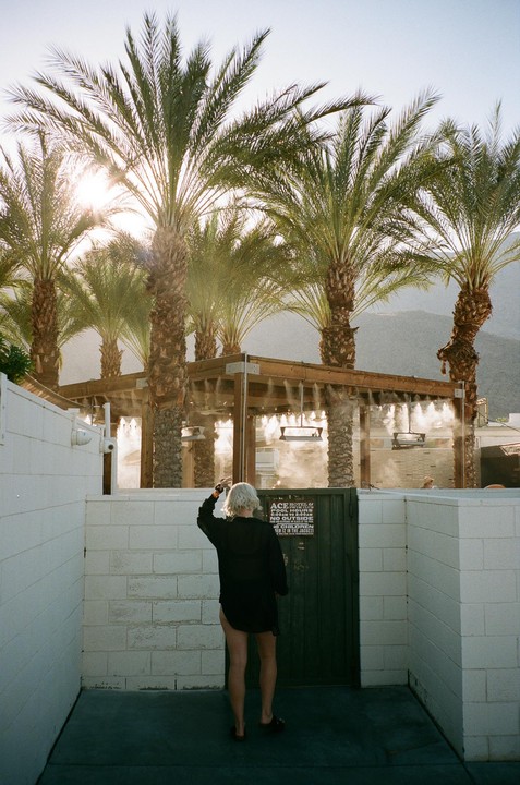 A woman stands in front of a gate with palm trees and a misting structure in the background.