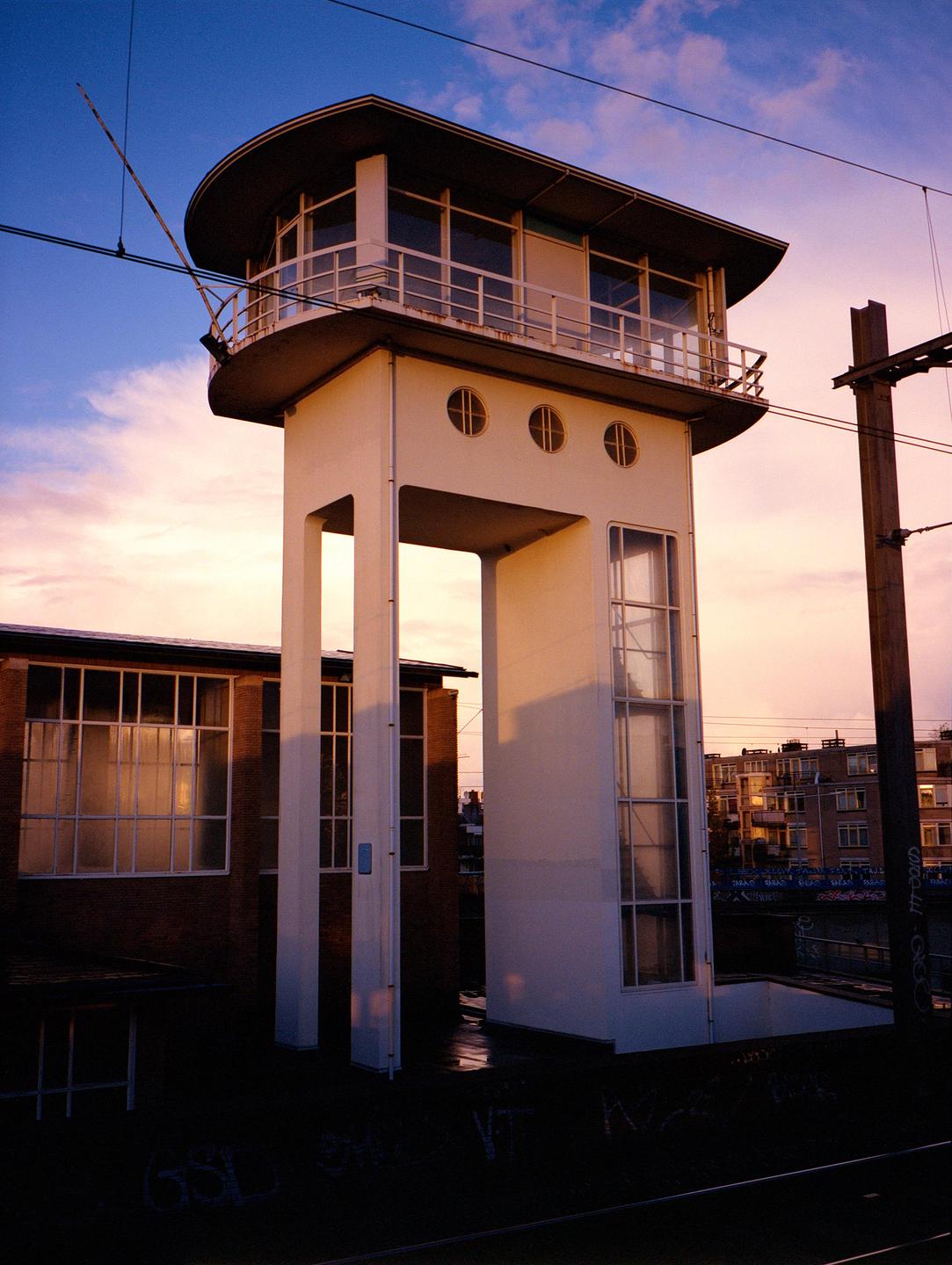 A tall, white, art deco building with a rounded top stands next to train tracks under a colorful sky.