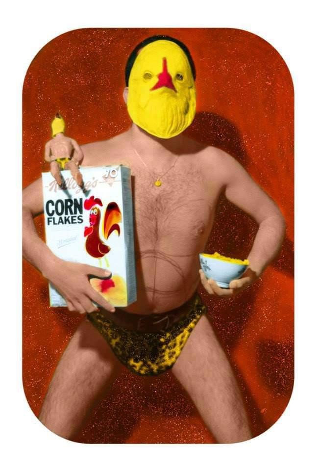 A man wearing a yellow bird mask and leopard print underwear holds a box of corn flakes and a bowl of cereal.