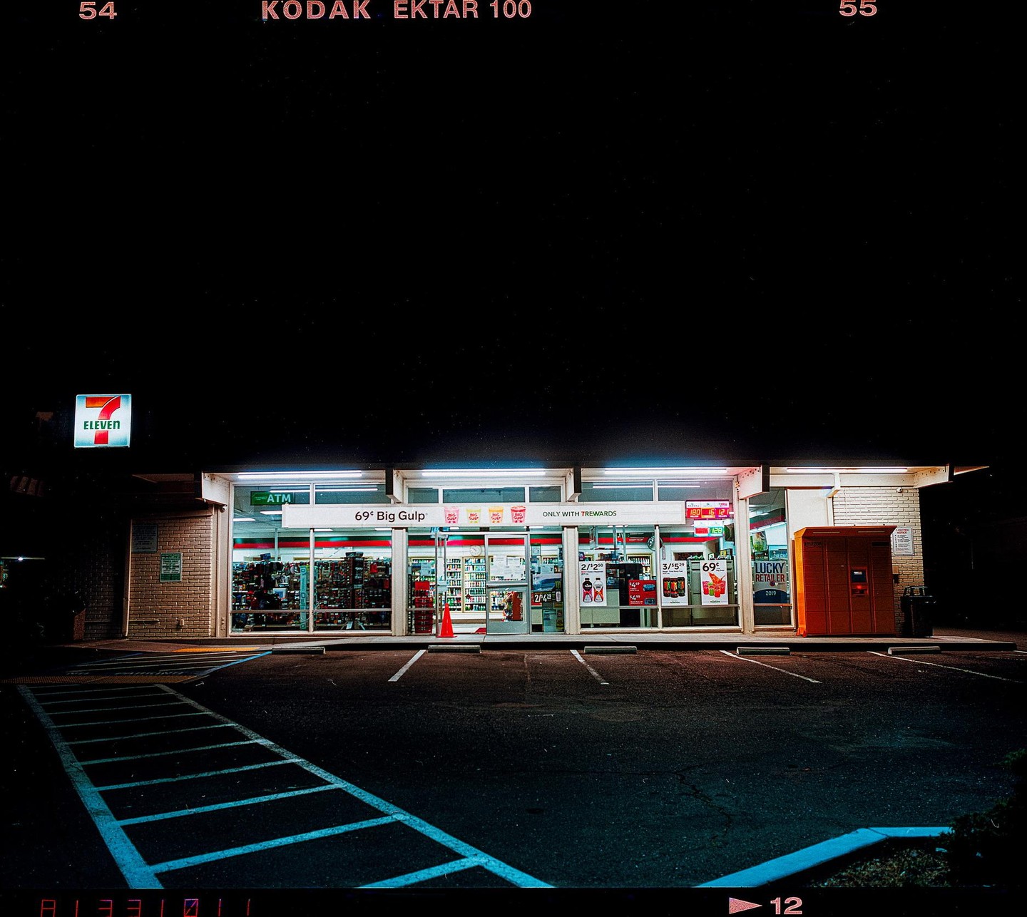 A brightly lit 7-Eleven convenience store stands at night with a dark parking lot in the foreground.