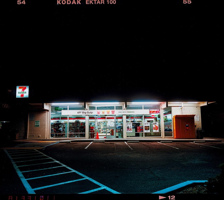 A brightly lit 7-Eleven convenience store stands at night with a dark parking lot in the foreground.