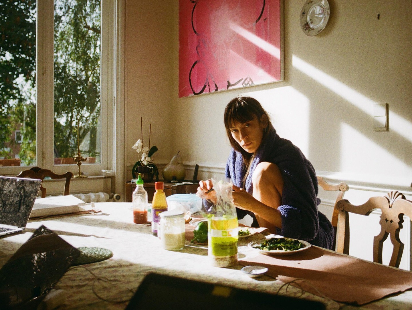 A woman in a purple robe sits at a table with food and a laptop, illuminated by sunlight streaming through a window.