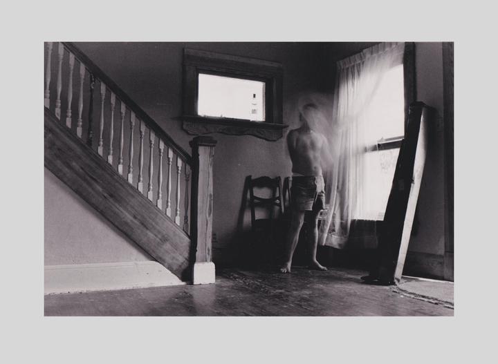 A shirtless man stands barefoot in front of a window with sheer curtains, next to a wooden staircase.