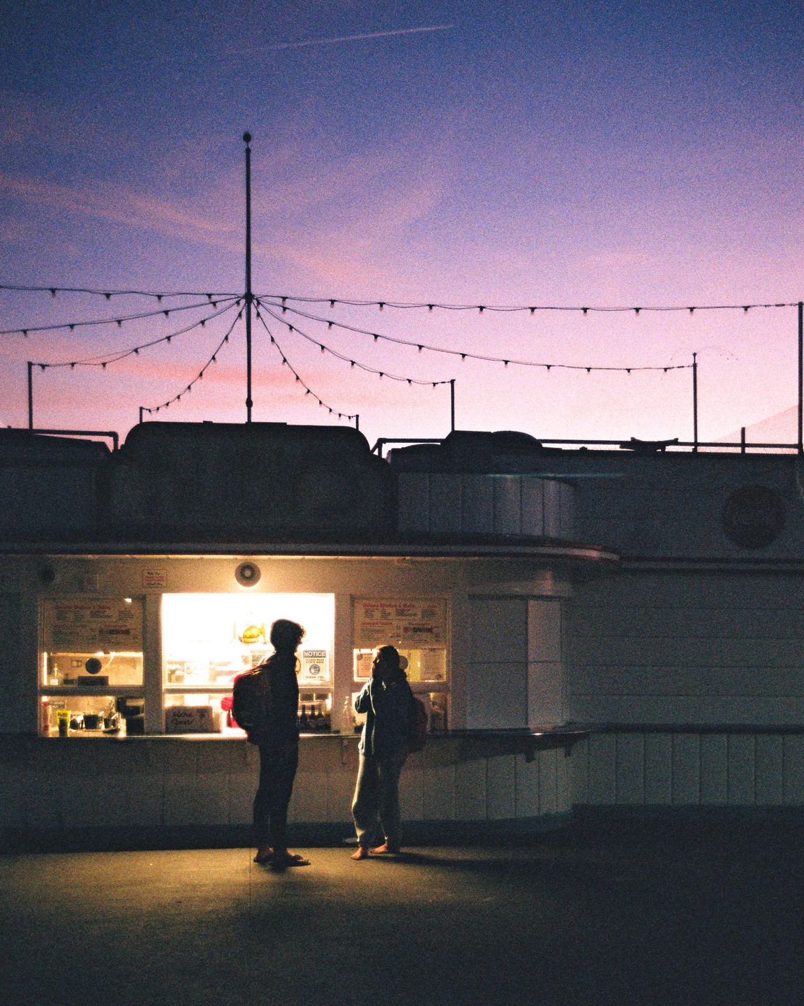 Two people stand in front of a brightly lit food stall at dusk.