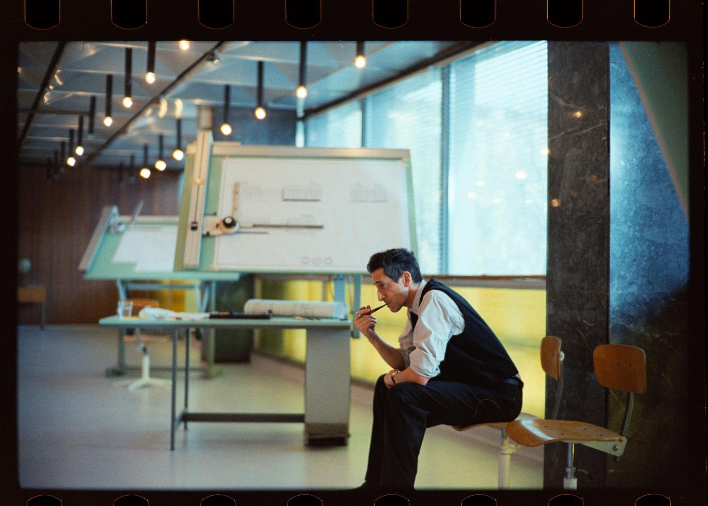 A man sits on a stool in an office with drafting tables and large windows, holding a pipe to his mouth.