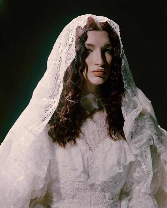 A woman with long wavy hair wears a white lace dress and veil.
