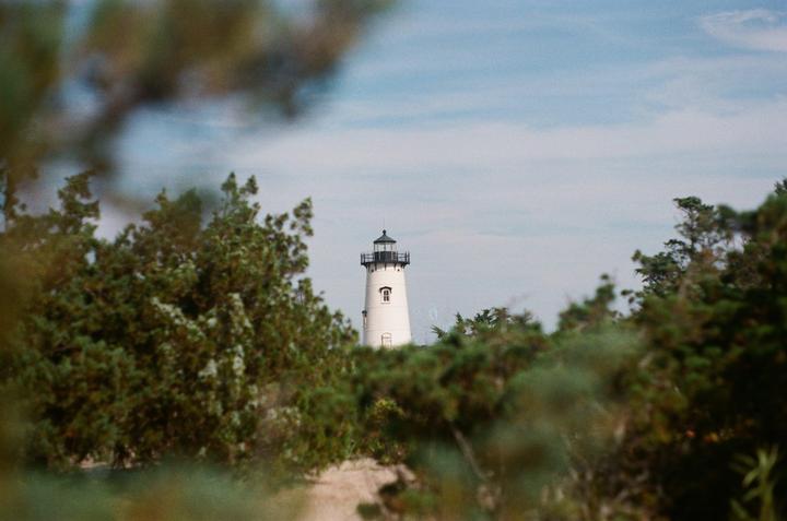 A white lighthouse with a black top is visible through green foliage under a cloudy sky.