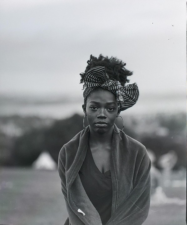 A woman wearing a head wrap and large hoop earrings looks directly at the viewer.