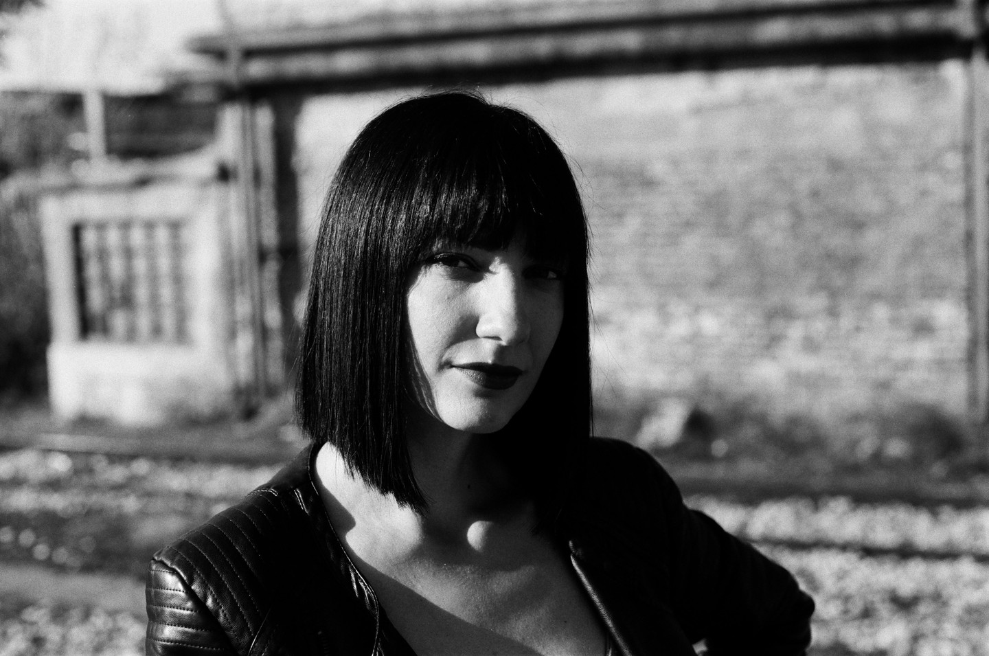 A woman with dark hair and a leather jacket looks at the camera in front of a brick wall.
