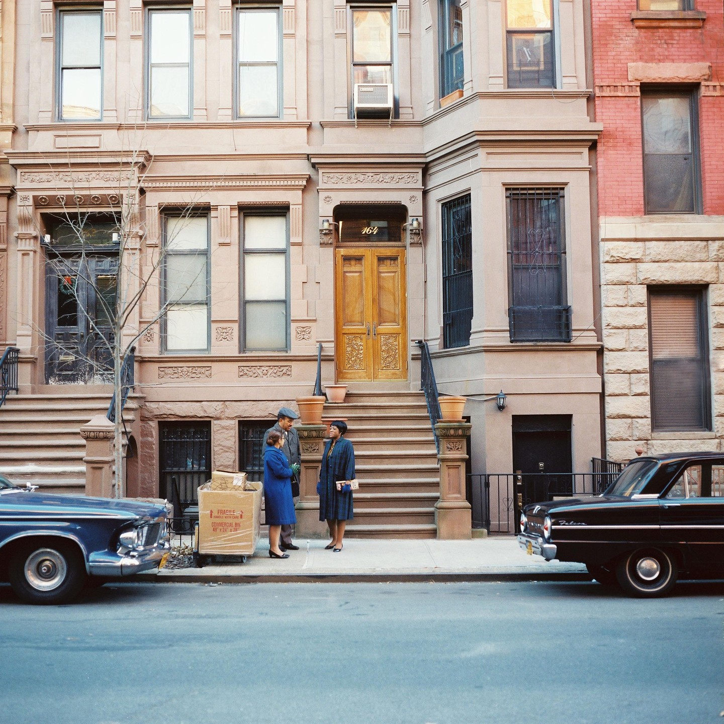 Two women and a man stand on a sidewalk next to a moving box, with two cars parked on the street and a brownstone building in the background.