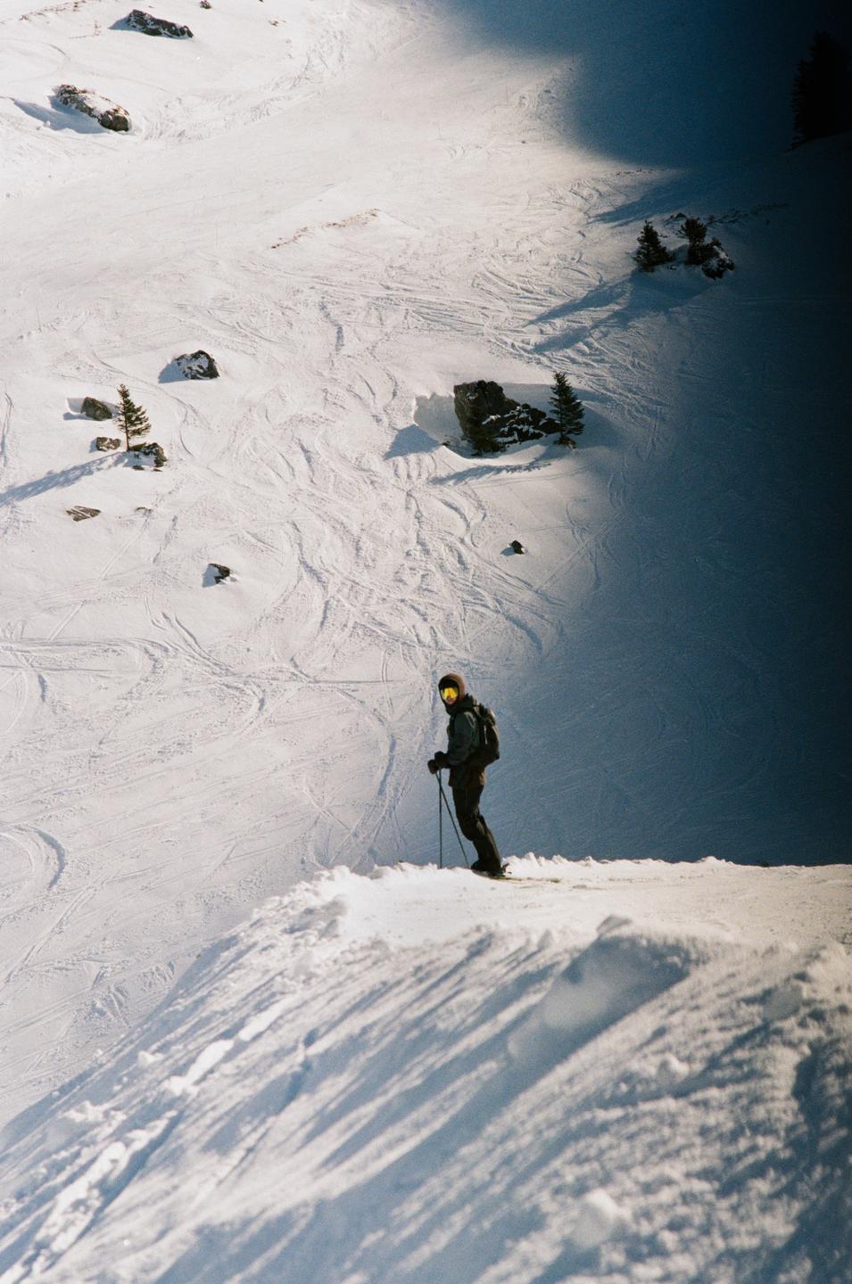 A skier stands on a snowy slope with ski tracks, rocks, and small trees visible in the background.