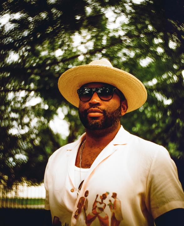 A man wearing a straw hat and sunglasses looks directly at the viewer against a blurred green background.